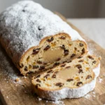 Ein ganzer Christstollen mit Puderzucker auf einem Holzbrett, daneben liegt eine aufgeschnittene Scheibe.