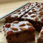 Mehrere quadratische Stücke Gewürzkuchen mit Schokoguss, Mandeln und Kirschen auf einer Kuchenplatte.