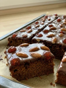 Mehrere quadratische Stücke Gewürzkuchen mit Schokoguss, Mandeln und Kirschen auf einer Kuchenplatte.