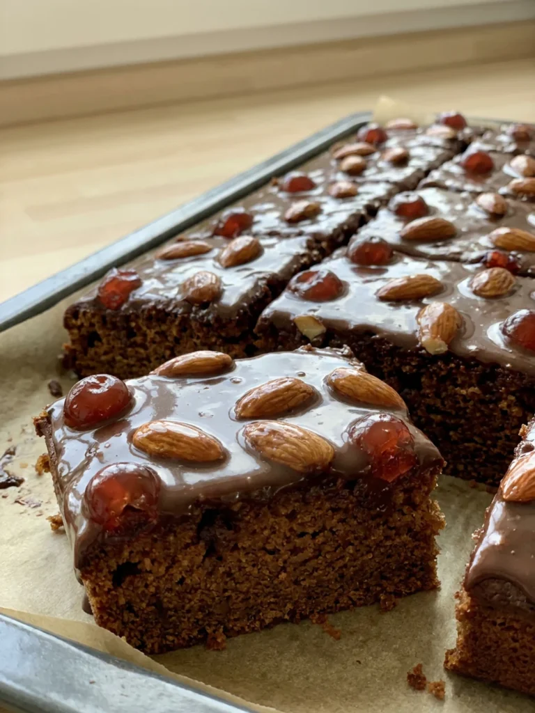 Mehrere quadratische Stücke Gewürzkuchen mit Schokoguss, Mandeln und Kirschen auf einer Kuchenplatte.
