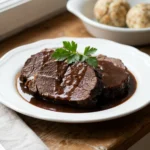 Ein Teller mit aufgeschnittenem Rinderbraten, übergossen mit glänzender Soße, daneben Kartoffelklöße.
